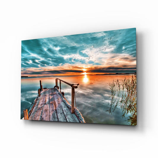 Wooden jetty with rope railing extending into calm water at sunset, with turquoise sky, flying birds, and golden sun reflection.