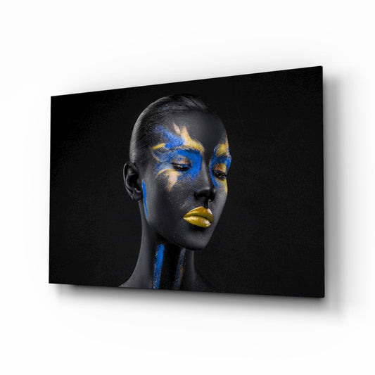 Woman's profile with black skin and blue and gold geometric paint patterns on face and neck against dark background on glass wall art.