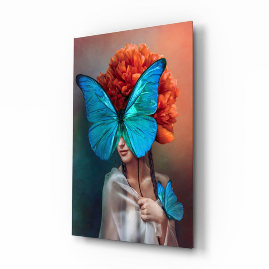 Glass wall art panel displayed at an angle showing a woman wearing a large blue butterfly mask and orange peony headdress.