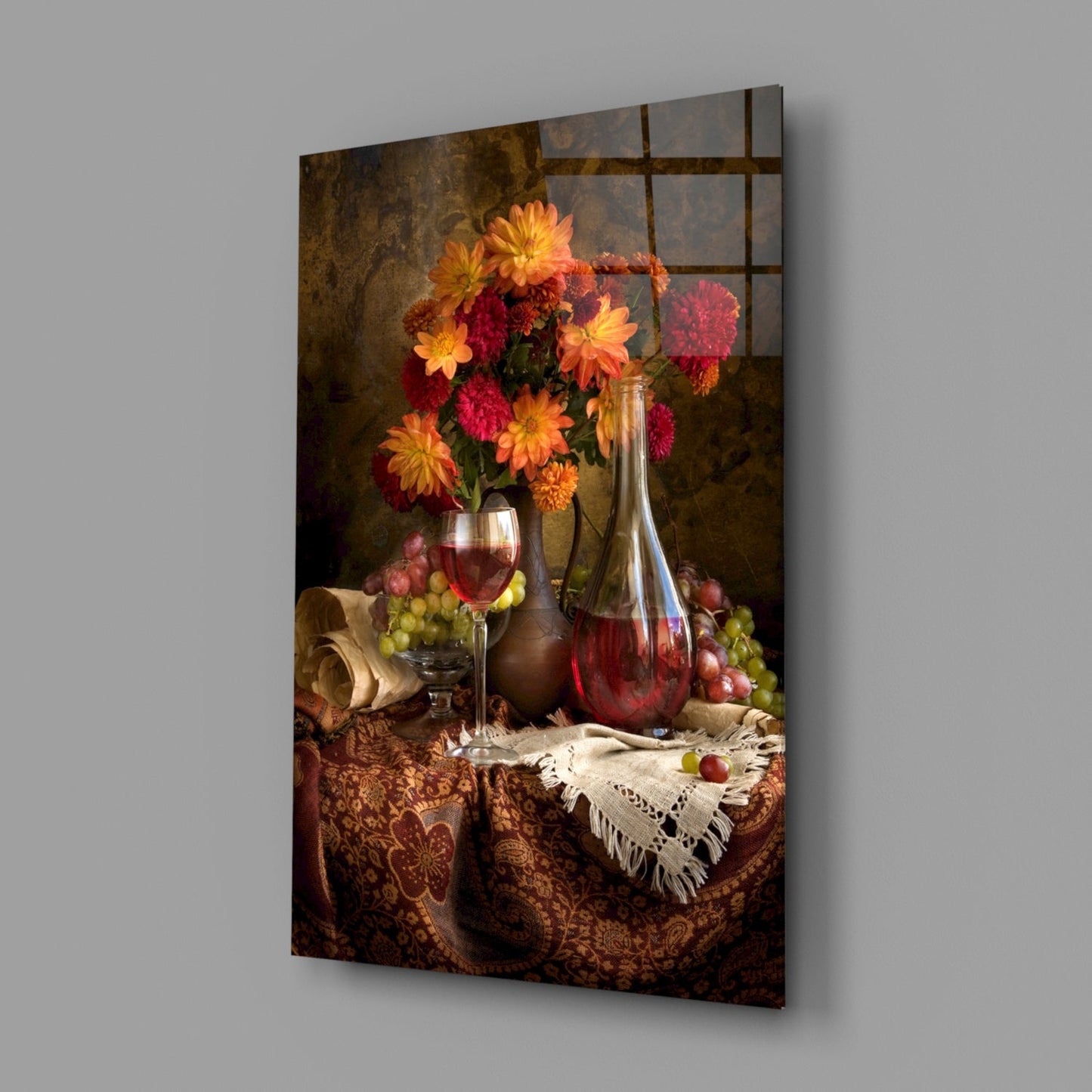 Glass wall art displaying a still life arrangement with red and yellow flowers in a vase, wine glasses, bottle, and grapes on a patterned brown fabric table.