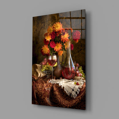 Glass wall art displaying a still life arrangement with red and yellow flowers in a vase, wine glasses, bottle, and grapes on a patterned brown fabric table.