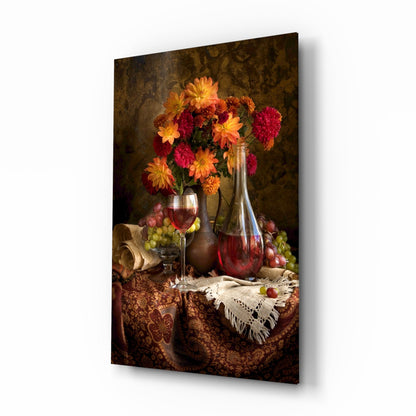 Glass wall art panel displaying a still life arrangement with red and yellow flowers in a vase, wine glass, bottle, and grapes on a patterned fabric-draped table against a textured brown background.