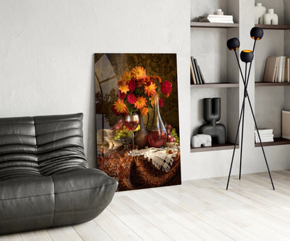 Glass wall art depicting red and yellow flowers in vases with wine glasses and fruit on draped fabric, displayed in modern living room setting.
