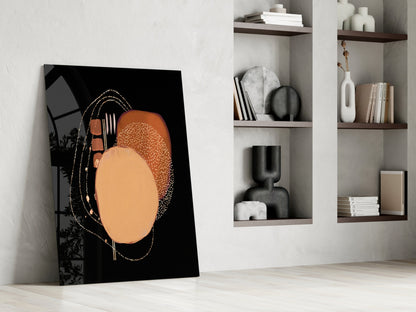 Abstract black glass wall art with orange and tan circular design leaning against white wall beside shelving.