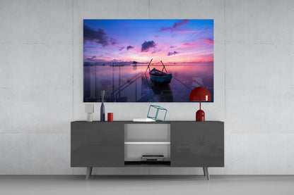 Glass wall art depicting a purple and pink sunset with a moored boat, displayed above a gray media console in a modern interior setting.
