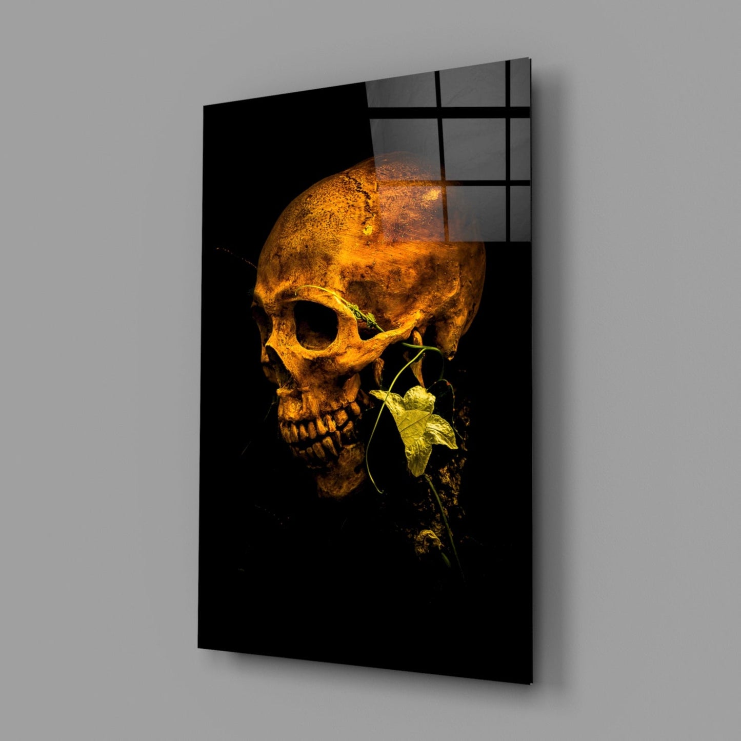 Gold-toned skull with yellow-green flower stem against black background, displayed as angled glass wall panel with grid pattern overlay.