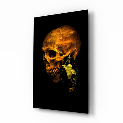 Golden skull with green flowering vine emerging from jaw against black background on glass panel, displayed at angle.