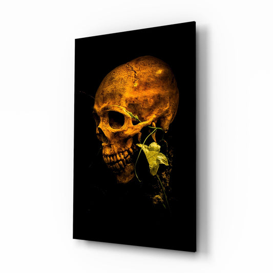 Golden skull with green flowering vine emerging from jaw against black background on glass panel, displayed at angle.