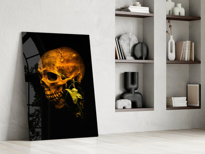 Golden skull with yellow flower on black background glass art leaning against white wall beside shelving.