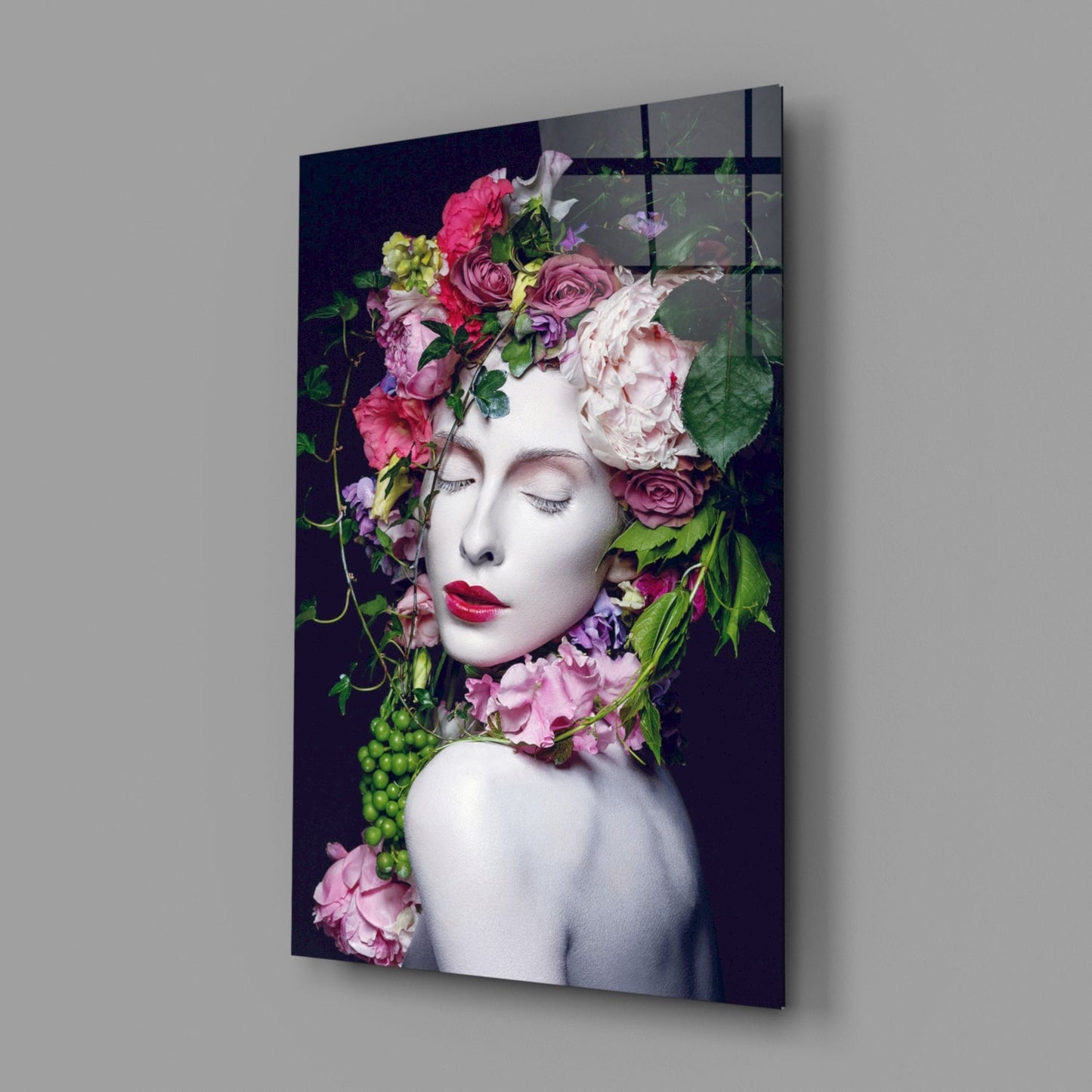 Angled view of glass wall art showing white sculptural face adorned with vibrant pink, red, and cream flowers against dark background.