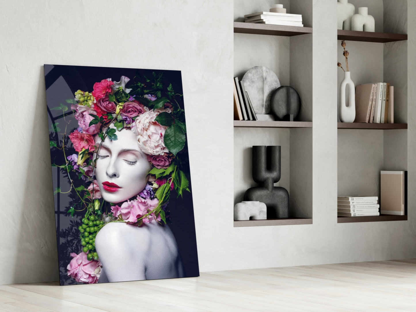 Glass wall art leaning against white wall displays woman's face with vibrant pink and purple flowers, dark background, styled interior setting with wooden shelves.