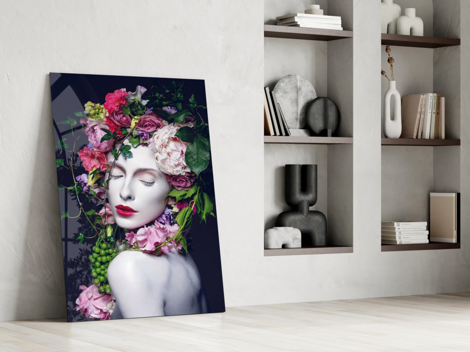 Glass wall art leaning against white wall displays woman's face with vibrant pink and purple flowers, dark background, styled interior setting with wooden shelves.