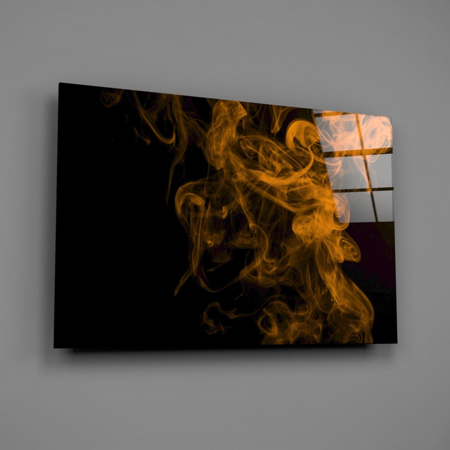 Glass wall art panel displaying golden-orange smoke swirls against black background, mounted at angle on gray wall.