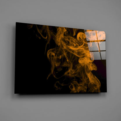 Glass wall art panel displaying golden-orange smoke swirls against black background, mounted at angle on gray wall.