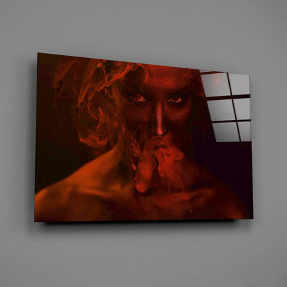 Glass wall art panel displaying a woman's face in red-orange tones surrounded by swirling smoke effects, mounted at an angle against a gray wall.