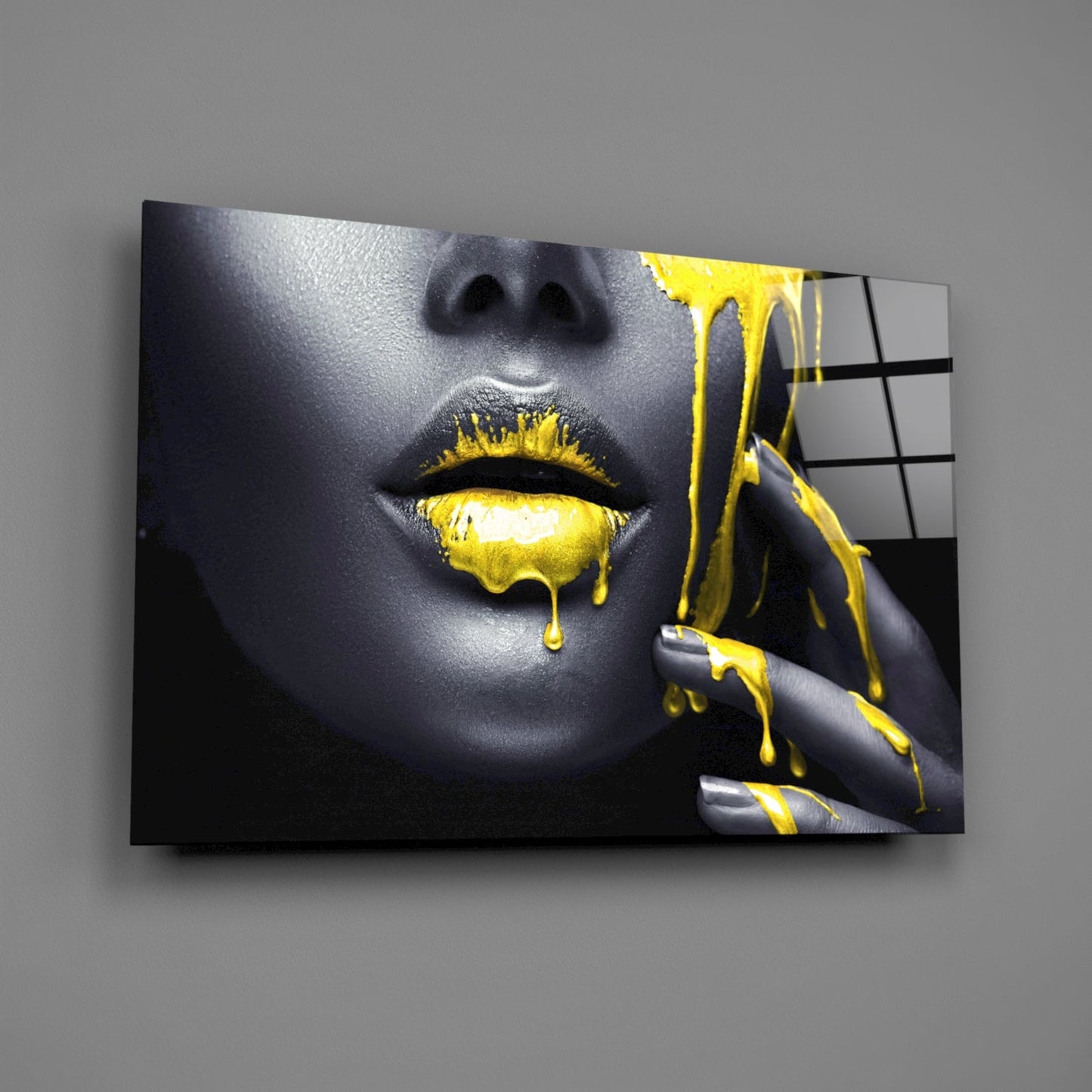 Glass wall art displaying a close-up of a woman's face with gray skin, yellow dripping lips and nails, mounted on gray wall.
