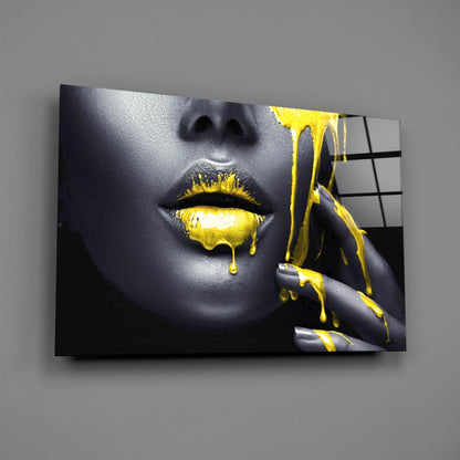 Glass wall art displaying a close-up of a woman's face with gray skin, yellow dripping lips and nails, mounted on gray wall.