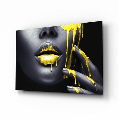 Glass wall art displaying a woman's face with gray skin and bright yellow paint dripping from lips and fingers against black background.
