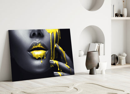 Black and gold glass wall art featuring a woman's face with yellow paint dripping from lips, leaning against white interior wall.