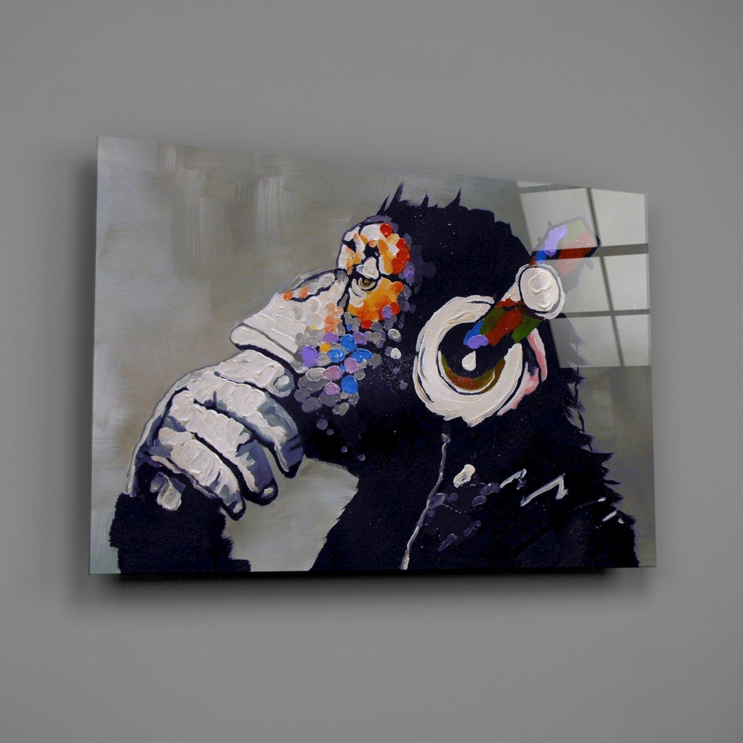 Glass wall art depicting a black chimpanzee wearing colorful headphones with vibrant paint splotches, mounted against a light wall.