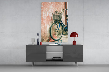 Vertical glass wall art depicting a blue bicycle with basket leaning against weathered terracotta wall, displayed above modern gray sideboard with red decorative objects.