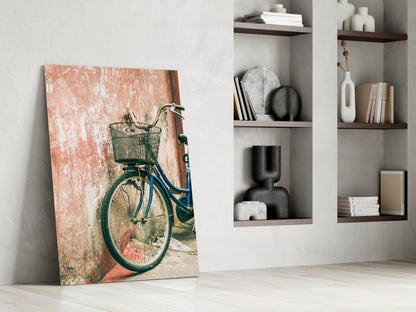 Canvas print of blue bicycle with basket leaning against weathered terracotta wall, displayed in modern room with white shelving.