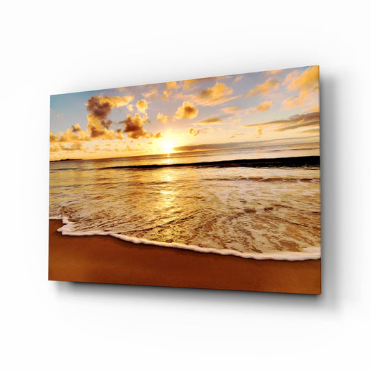 Glass wall art panel displaying a beach sunset with golden clouds, foamy waves, and sandy shore in warm amber tones.