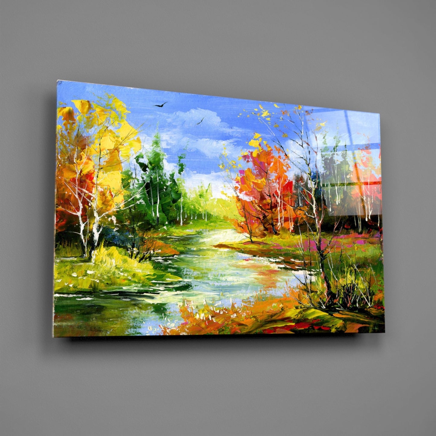 Vibrant autumn landscape glass wall art showing a stream, colorful fall foliage, and blue sky, displayed at an angle against gray wall.