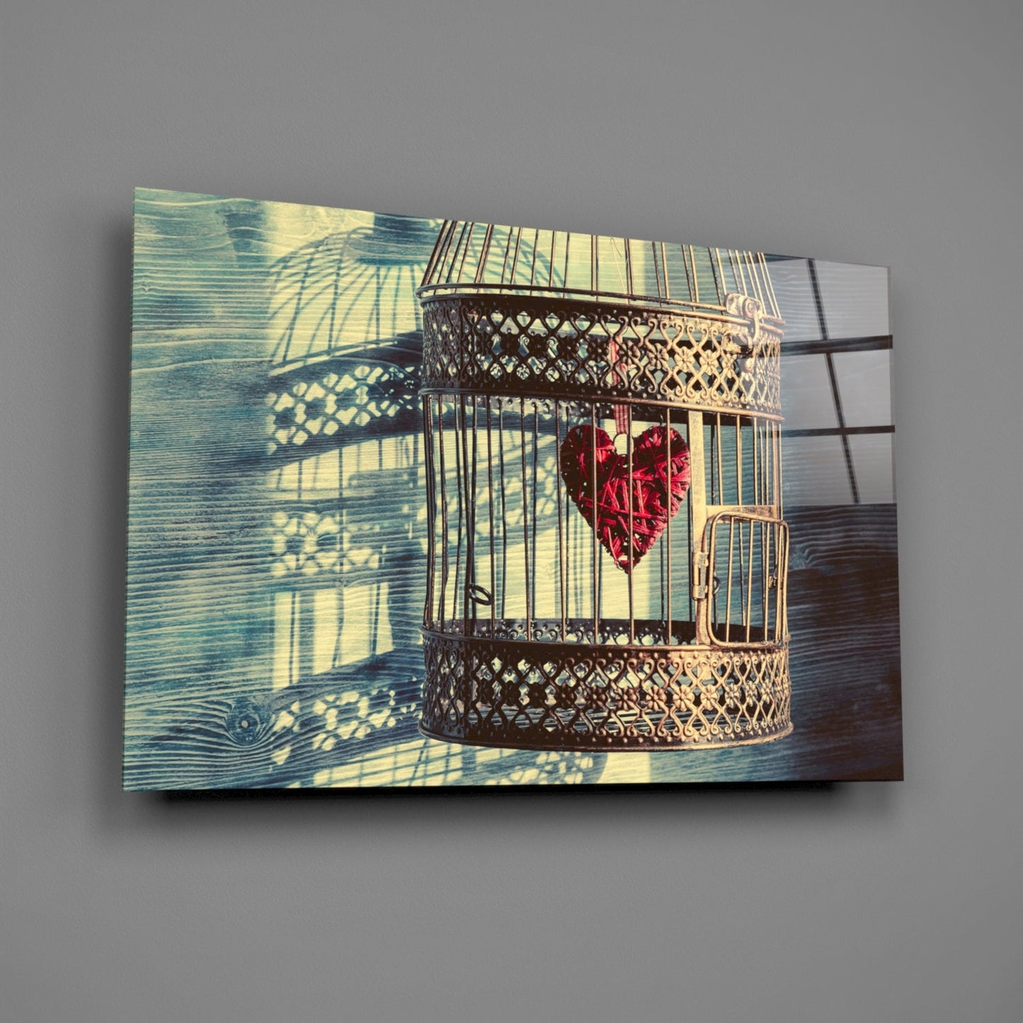 Decorative glass wall art depicting an ornate brass birdcage with red heart inside, casting geometric shadows on blue-green wooden background.
