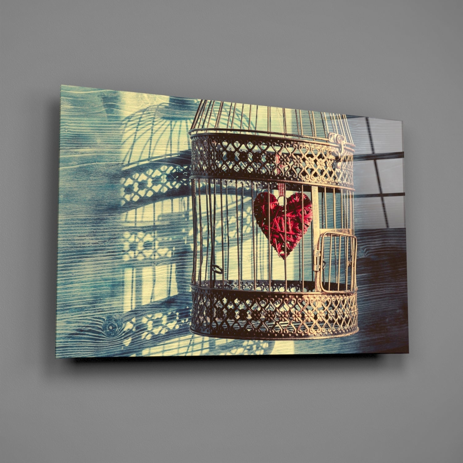 Decorative glass wall art depicting an ornate brass birdcage with red heart inside, casting geometric shadows on blue-green wooden background.