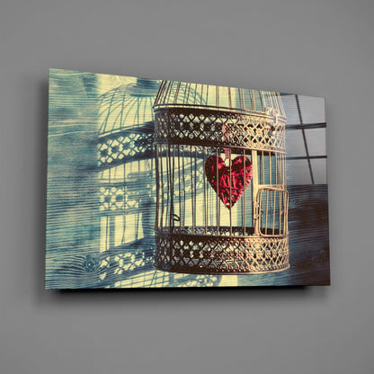 Decorative glass wall art depicting an ornate brass birdcage with red heart inside, casting geometric shadows on blue-green wooden background.