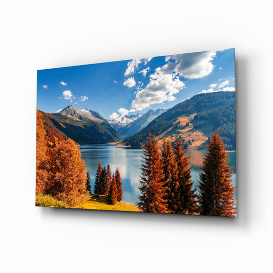 Angled view of glass wall art displaying an alpine lake surrounded by autumn-colored larch trees, mountains, and blue sky with clouds.