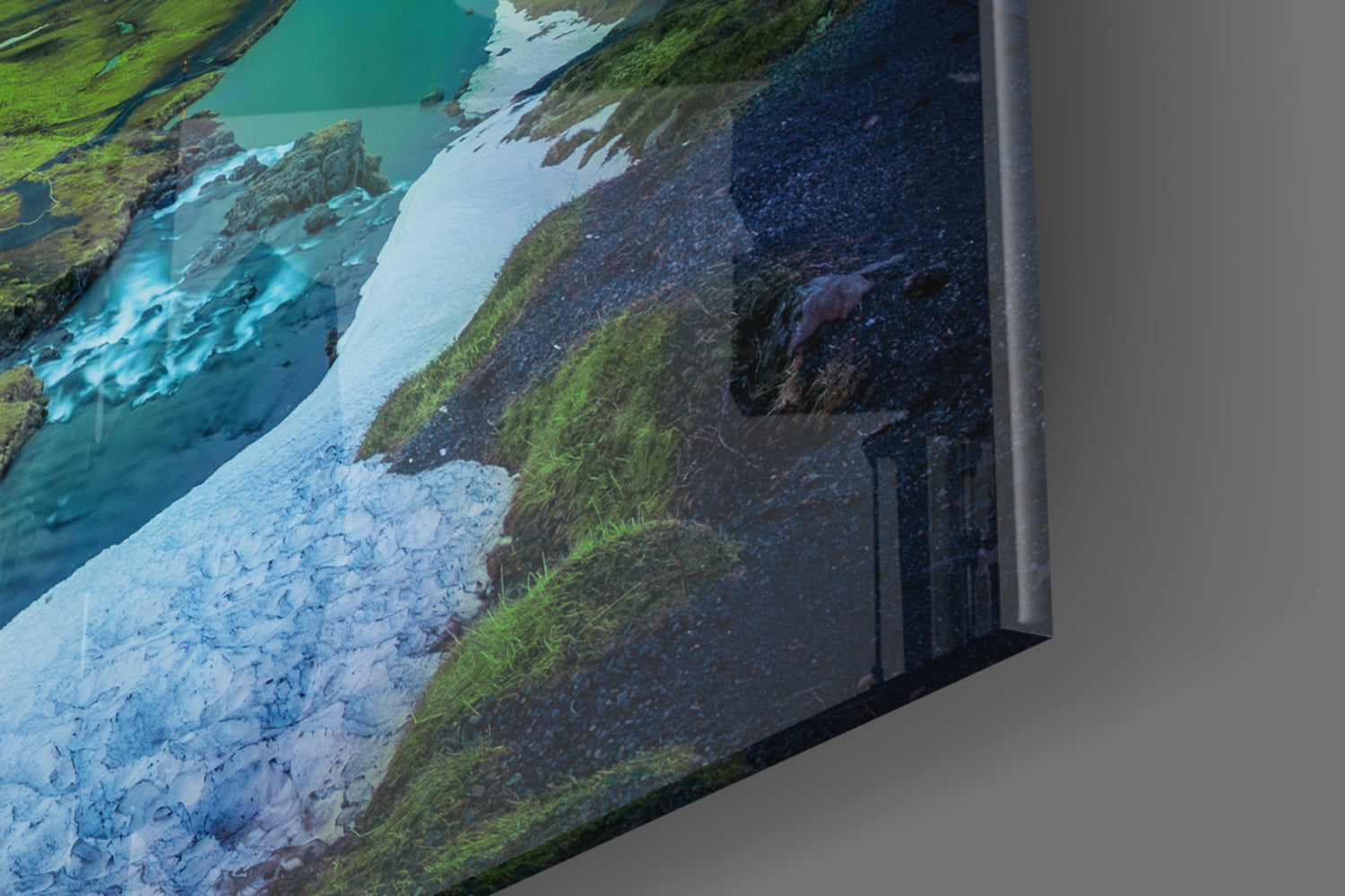 Angled view of glass wall art displaying a glacial river landscape with turquoise water, white glacier, green moss, and dark volcanic terrain.