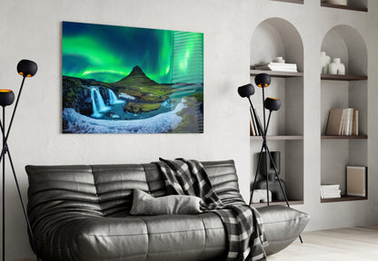 Northern Lights glass wall art displaying green aurora borealis over Icelandic waterfall and mountain landscape, hung above gray leather sofa in modern living room.