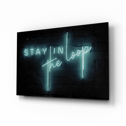 Cyan neon "Stay in the Loop" text glowing against dark brick wall on angled glass panel viewed from front-right perspective.