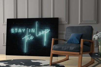 Cyan neon "Stay in the Loop" text glows on black glass panel displayed in modern living room with wooden chair and potted flowers.