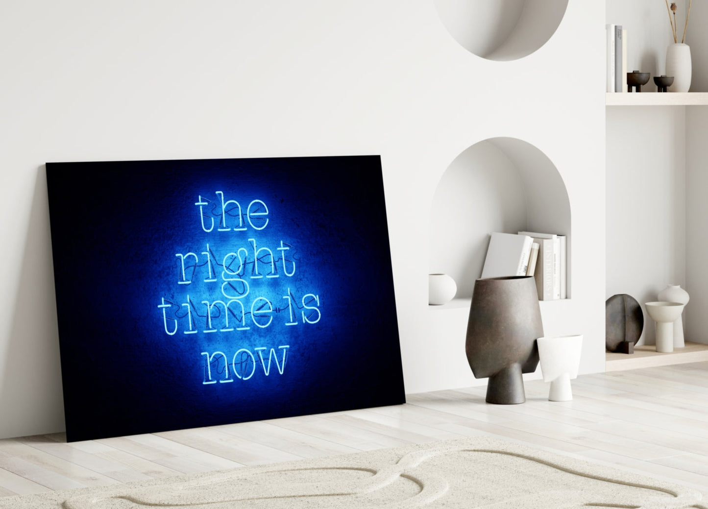 Blue neon "the right time is now" glass art leaning against white wall in minimalist room with shelving and decorative objects.