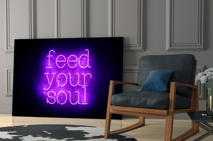 Purple neon "Feed Your Soul" glass wall art displayed on a dark screen next to a wooden-framed armchair in a modern interior setting.