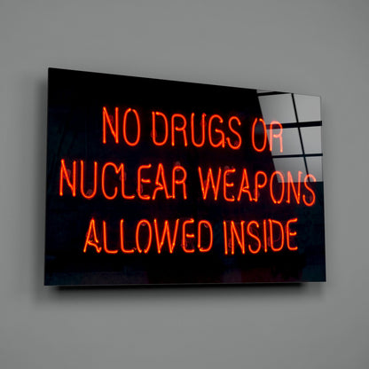 Neon sign on black glass displaying red glowing text reading "NO DRUGS OR NUCLEAR WEAPONS ALLOWED INSIDE" mounted on gray wall.