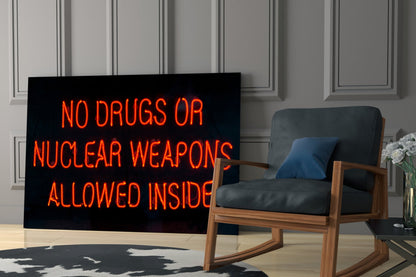 Red neon sign reading "NO DRUGS OR NUCLEAR WEAPONS ALLOWED INSIDE" displayed in modern living room with wooden chair and white flowers.