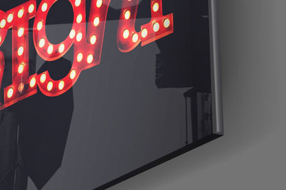 Red neon sign with illuminated bulbs mounted on dark glass wall, partial view of letters visible against grey wall.
