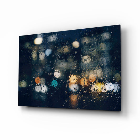 Angled perspective of glass wall art panel displaying rain droplets and blurred city lights in warm and cool tones against dark background.
