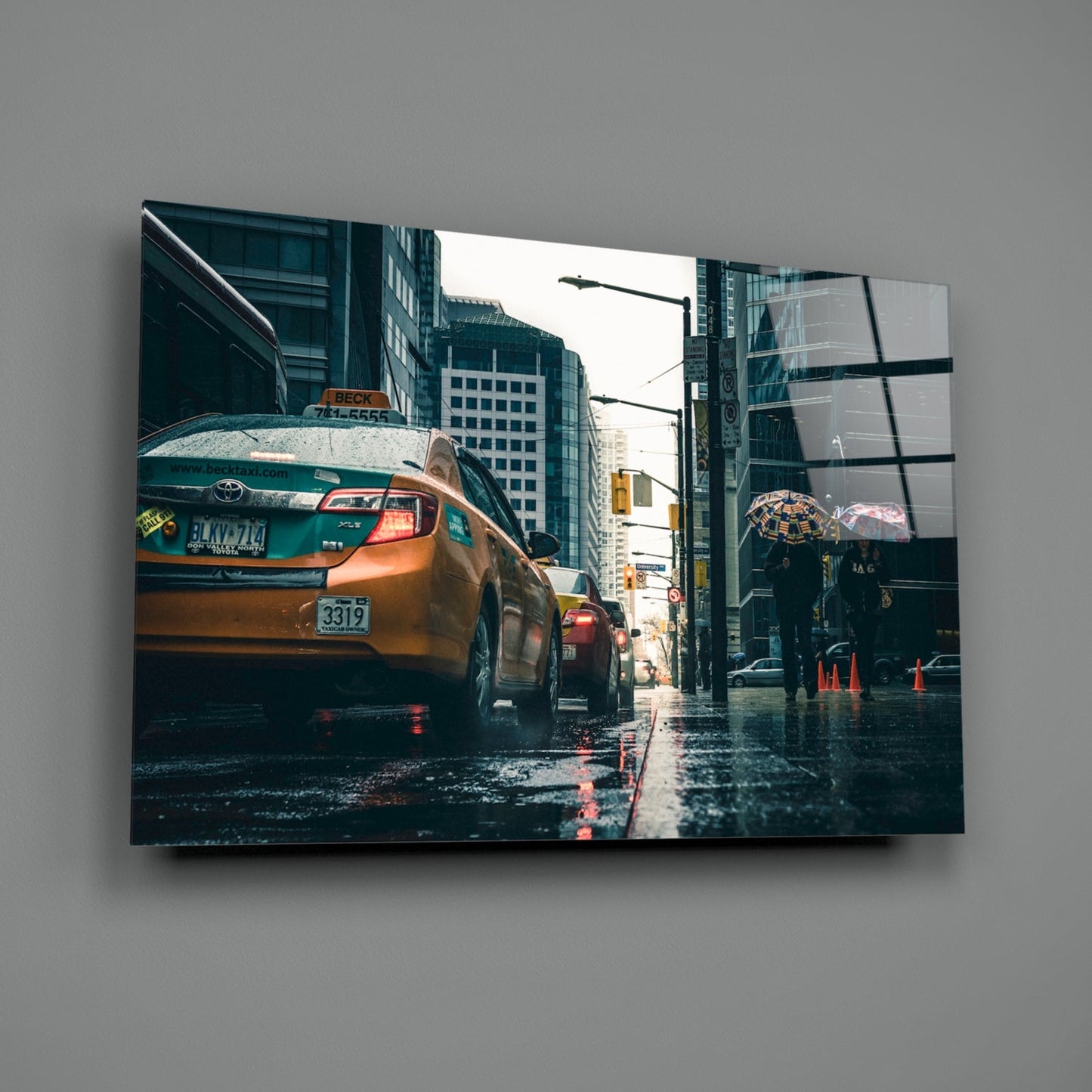 Glass wall art mounted on gray wall showing wet urban street with yellow taxi cab and rainy cityscape.