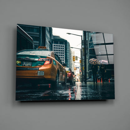 Glass wall art mounted on gray wall showing wet urban street with yellow taxi cab and rainy cityscape.