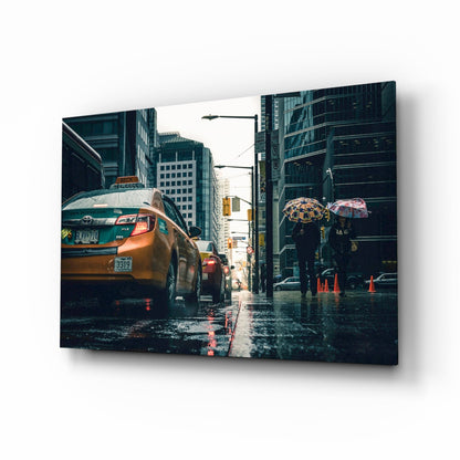 Glass wall art panel displayed at an angle showing a rainy New York City street with yellow taxi cab and pedestrians holding umbrellas.