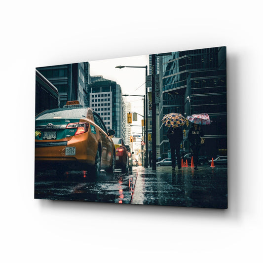 Glass wall art panel displayed at an angle showing a rainy New York City street with yellow taxi cab and pedestrians holding umbrellas.