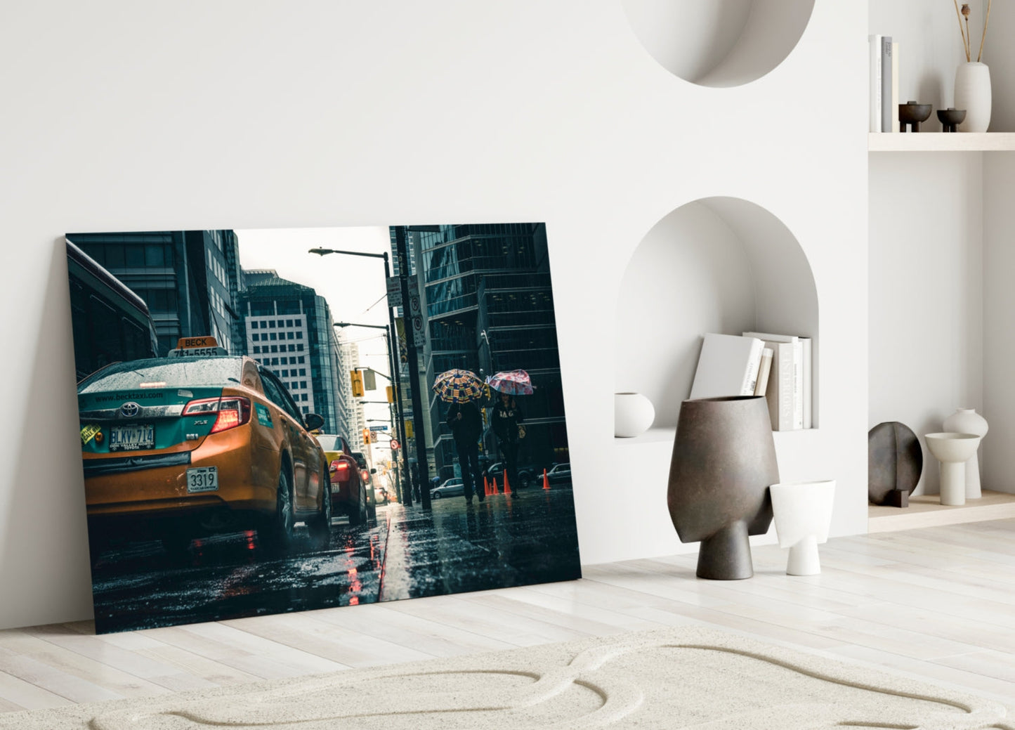 Leaning glass wall art featuring a wet urban street with a yellow taxi cab and pedestrians with umbrellas, displayed in a minimalist white interior.