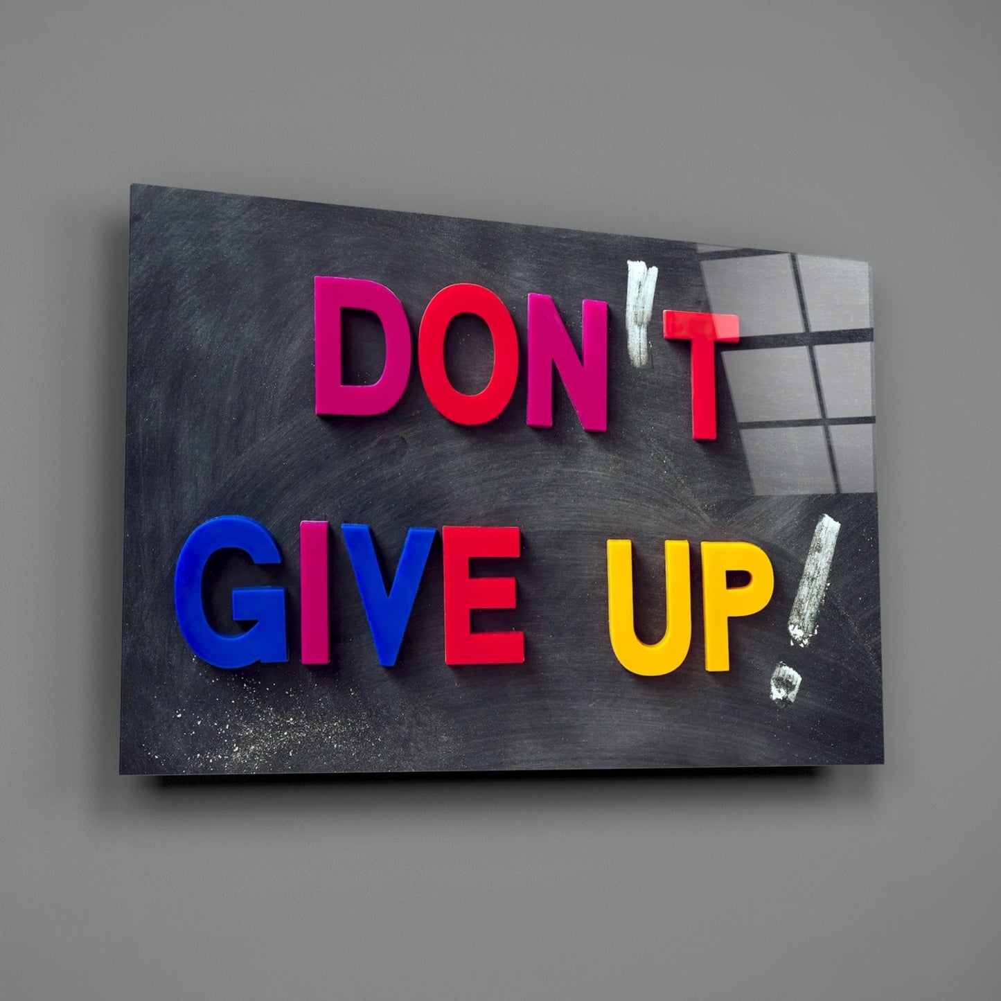 Colorful 3D letters spelling "DON'T GIVE UP" mounted on a dark textured glass panel against a gray wall.