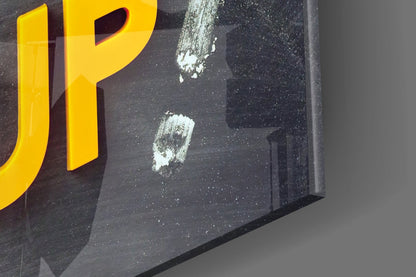 Close-up of black glass wall art with yellow "P" letter and white paint splatters visible on surface.