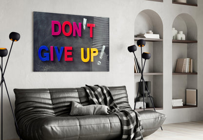 Colorful "Don't Give Up" glass wall art in pink, blue, and yellow text displayed above a black leather sofa in a modern living room interior.